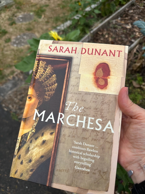 Outside, in a garden, a hand holds a book: The Marchesa, by Sarah Dunant. The cover includes a picture of Isabella d'Este.